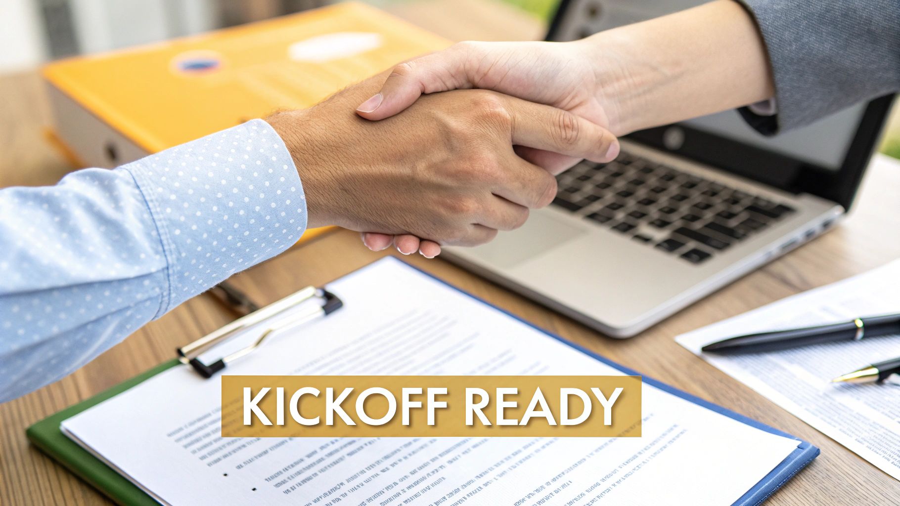 Two business professionals shake hands over documents, a laptop, and a binder, signifying a ready partnership.